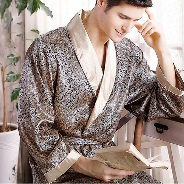Homme portant le peignoir Tanguy, un modèle tendance et agréable en satin brillant à motifs dorés, assis près d’une fenêtre et lisant un livre dans une ambiance cosy.