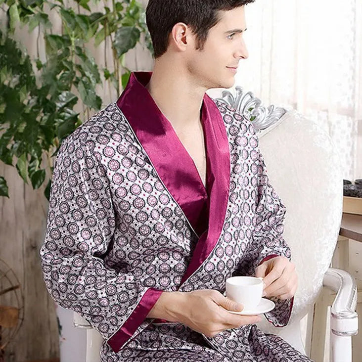Homme portant le peignoir Tanguy, un modèle tendance et agréable en satin imprimé avec col et manches bordeaux, assis dans un intérieur lumineux et tenant une tasse de café.