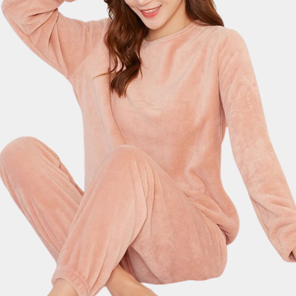 Femme assise, souriante, vêtue d’un pyjama polaire rose poudré, composé d’un haut à manches longues et d’un pantalon ample avec chevilles resserrées. Ensemble doux, léger et confortable, parfait pour des moments de détente à la maison.