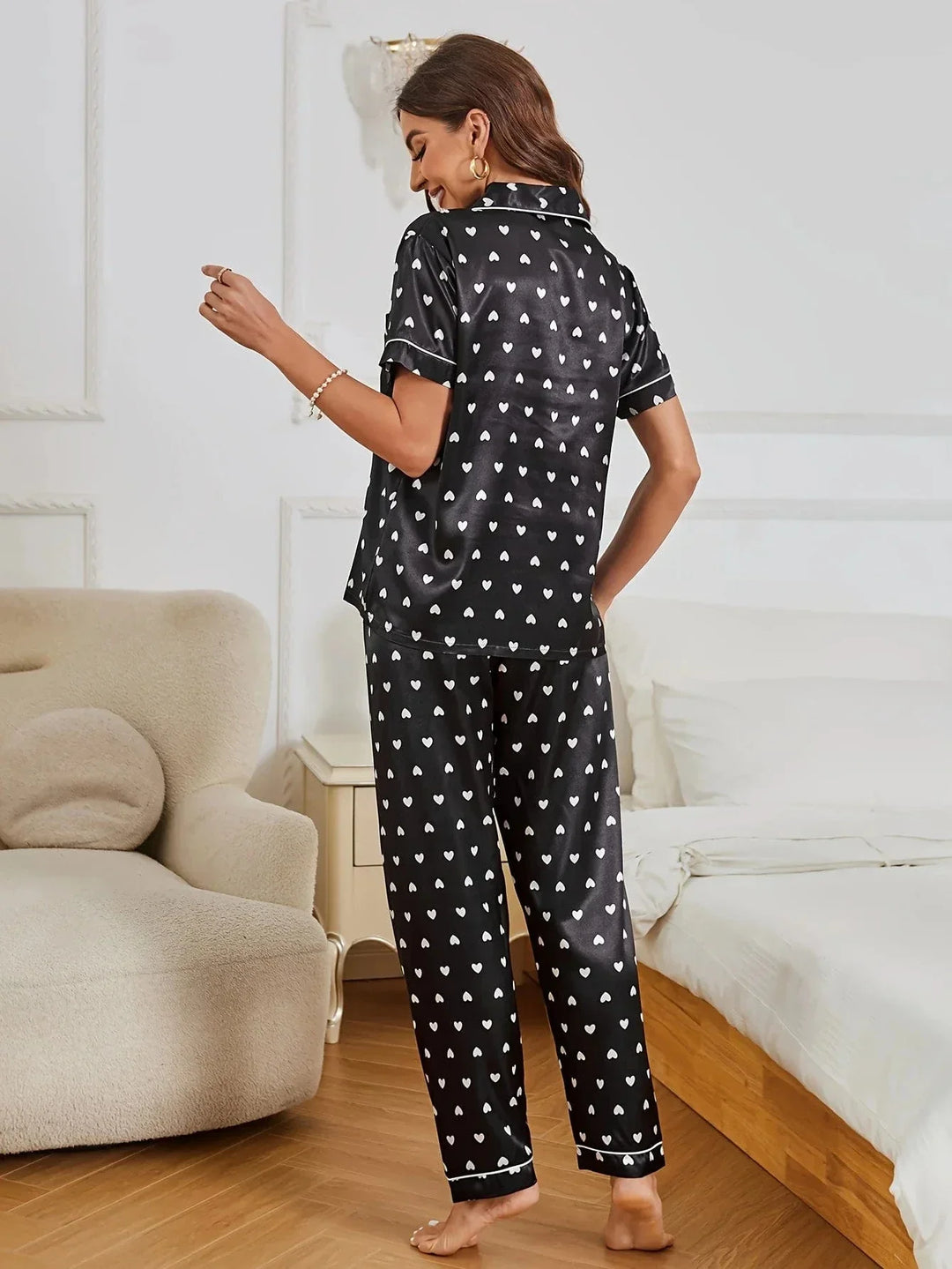 Pyjama en satin noir pour femme, avec une chemise décorée de petits cœurs blancs et un pantalon assorti. Sur cette photo, une femme porte ce pyjama élégant et romantique, avec une vue de dos, affichant un sourire détendu. Ce pyjama doux et confortable est parfait pour des moments de relaxation et de tranquillité à la maison.