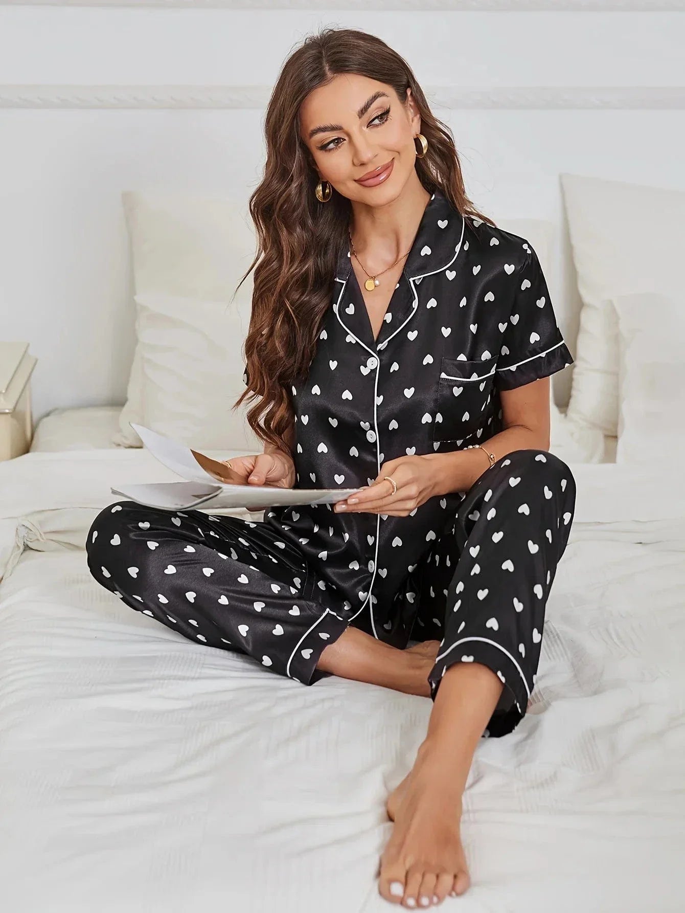 Pyjama en satin noir pour femme, avec une chemise et un pantalon décorés de petits cœurs blancs. Sur cette photo, une femme porte ce pyjama élégant et romantique, assise sur un lit avec un sourire radieux. Ce pyjama doux et confortable est idéal pour des moments de détente à la maison, alliant style et confort.