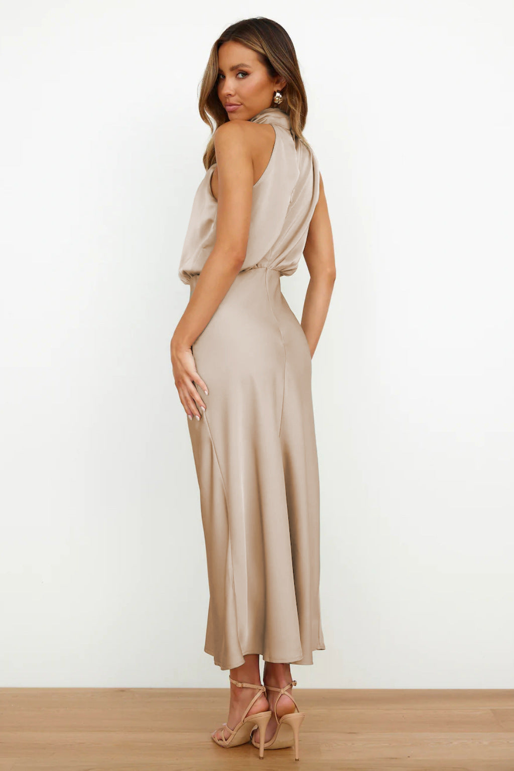 Vue de dos, cette robe longue beige en satin présente un design fluide et gracieux, avec un dos légèrement dégagé et un tissu qui épouse délicatement les courbes. Un choix idéal pour une allure chic et sophistiquée en été.