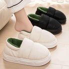 Chaussons unisexe Alex style doudoune, modèle blanc avec doublure verte porté à la maison, avec variante noire en arrière-plan, semelle épaisse antidérapante et intérieur chaud.