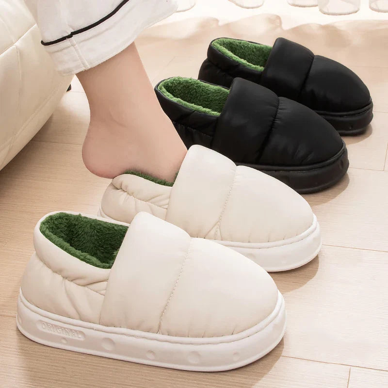 Chaussons unisexe Alex style doudoune, modèle blanc avec doublure verte porté à la maison, avec variante noire en arrière-plan, semelle épaisse antidérapante et intérieur chaud.