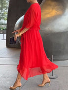 Femme marchant dans un espace moderne, portant une robe longue et fluide de couleur rouge, avec des accessoires dorés et des talons transparents.