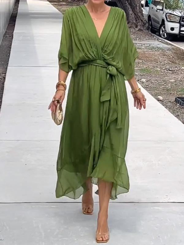 Femme portant une robe longue en chiffon vert olive, cintrée à la taille avec un nœud, et accompagnée d'un sac à main doré et des bracelets dorés.