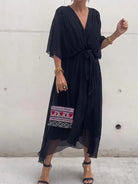 Femme portant une robe noire en chiffon fluide, avec une taille cintrée et un décolleté en V, accompagnée d'un sac à main coloré et des accessoires dorés.