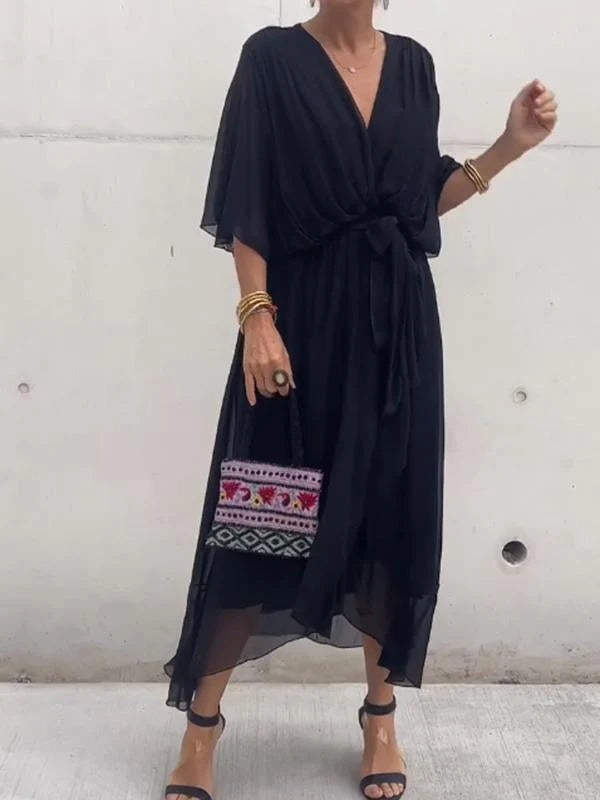 Femme portant une robe noire en chiffon fluide, avec une taille cintrée et un décolleté en V, accompagnée d'un sac à main coloré et des accessoires dorés.