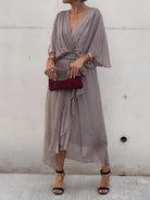 Femme portant une robe en chiffon taupe, fluide et légère, avec un décolleté en V et une ceinture à nouer à la taille. Elle accessoirise la robe avec un sac à main bordeaux et des sandales noires.