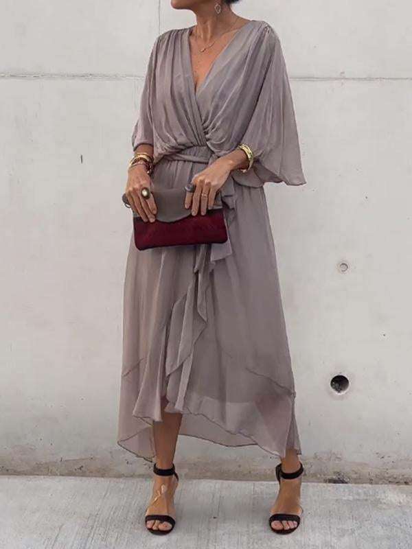 Femme portant une robe en chiffon taupe, fluide et légère, avec un décolleté en V et une ceinture à nouer à la taille. Elle accessoirise la robe avec un sac à main bordeaux et des sandales noires.