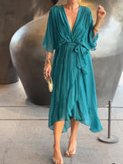 Femme portant une robe chiffon turquoise fluide avec un décolleté en V et des manches larges. Elle accessoirise la tenue avec une pochette dorée et des bracelets dorés.
