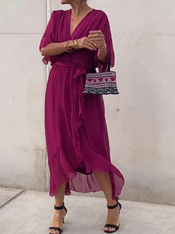 Femme portant une robe chiffon fuchsia avec des manches fluides et un décolleté en V. Elle accessoirise la tenue avec un sac à main tissé et des sandales noires à talons.