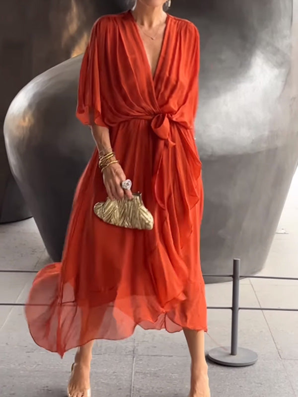 Femme portant une robe chiffon rouge vif, avec des manches amples et un décolleté en V, accessoirisée d'une pochette dorée et des bracelets dorés.