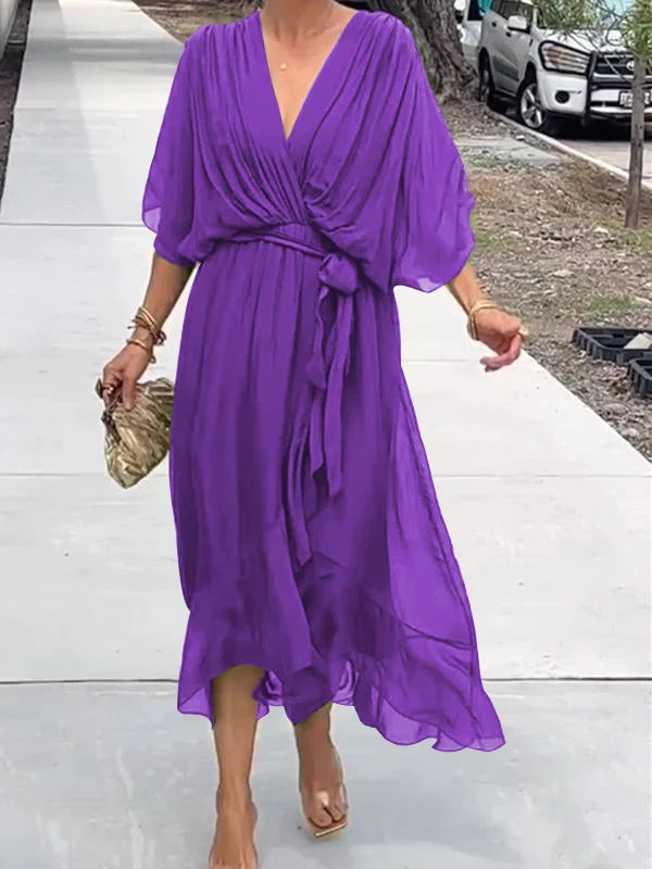 Femme marchant dans une robe chiffon violette, avec des manches larges et une ceinture assortie, portant une pochette dorée et des sandales transparentes.