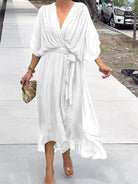 Femme marchant dans une robe chiffon blanche, avec des manches larges et une ceinture assortie, portant une pochette dorée et des sandales transparentes.