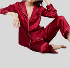 Femme assise portant un pyjama deux pièces en satin rouge, composé d’un haut à manches longues boutonné avec col à revers et passepoils blancs, et d’un pantalon fluide assorti. Ensemble raffiné, doux et élégant, idéal pour passer des nuits confortables avec style.