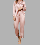 Femme portant un pyjama deux pièces en satin rose poudré, composé d’un haut boutonné à col classique et passepoils fins, et d’un pantalon long fluide avec taille élastique. Ensemble raffiné, soyeux et confortable, idéal pour des nuits élégantes et relaxantes.