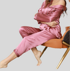Femme assise sur une chaise design, vêtue d’un pyjama deux pièces en satin rose ancien avec passepoils blancs, comprenant un haut boutonné à col classique et un pantalon fluide assorti. Ensemble chic, doux et confortable, parfait pour les soirées cocooning et les nuits élégantes.