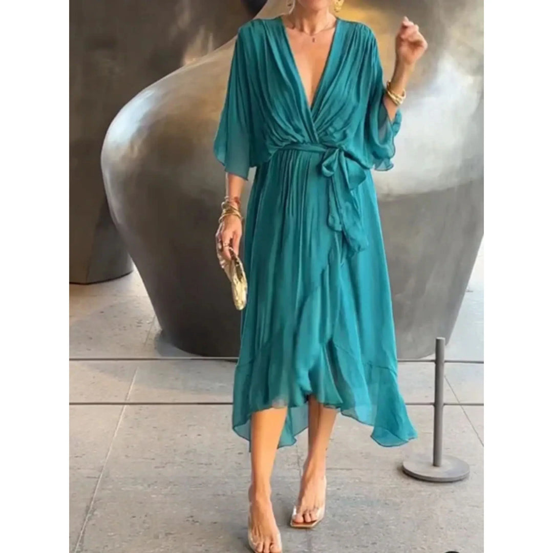 Vert turquoise fluide, décolleté cache-cœur, manches amples et ceinture nouée à la taille, longueur midi avec ourlet volanté, tenant une pochette.