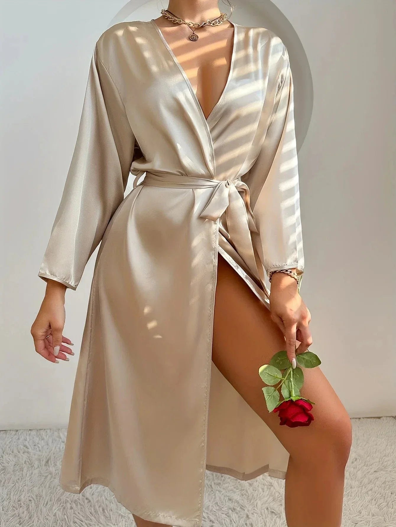 Femme debout tenant une rose rouge, vêtue d’un peignoir satiné couleur champagne, avec col croisé, ceinture à nouer et fente élégante sur la jambe. Modèle raffiné et fluide, parfait pour une allure romantique et confortable à la maison.
