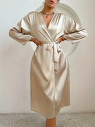 Femme debout, mains sur les hanches, portant un peignoir satiné couleur champagne à col croisé, manches longues et ceinture nouée à la taille. Modèle fluide, sobre et élégant, idéal pour une tenue de détente chic à la maison ou en sortie de lit raffinée.