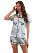 Femme portant un pyjama court en satin gris clair, composé d’un haut à manches courtes boutonné avec passepoil noir et d’un short assorti. Motifs papillons bleus imprimés sur l’ensemble. Pyjama doux, fluide et élégant, parfait pour les nuits confortables avec style.