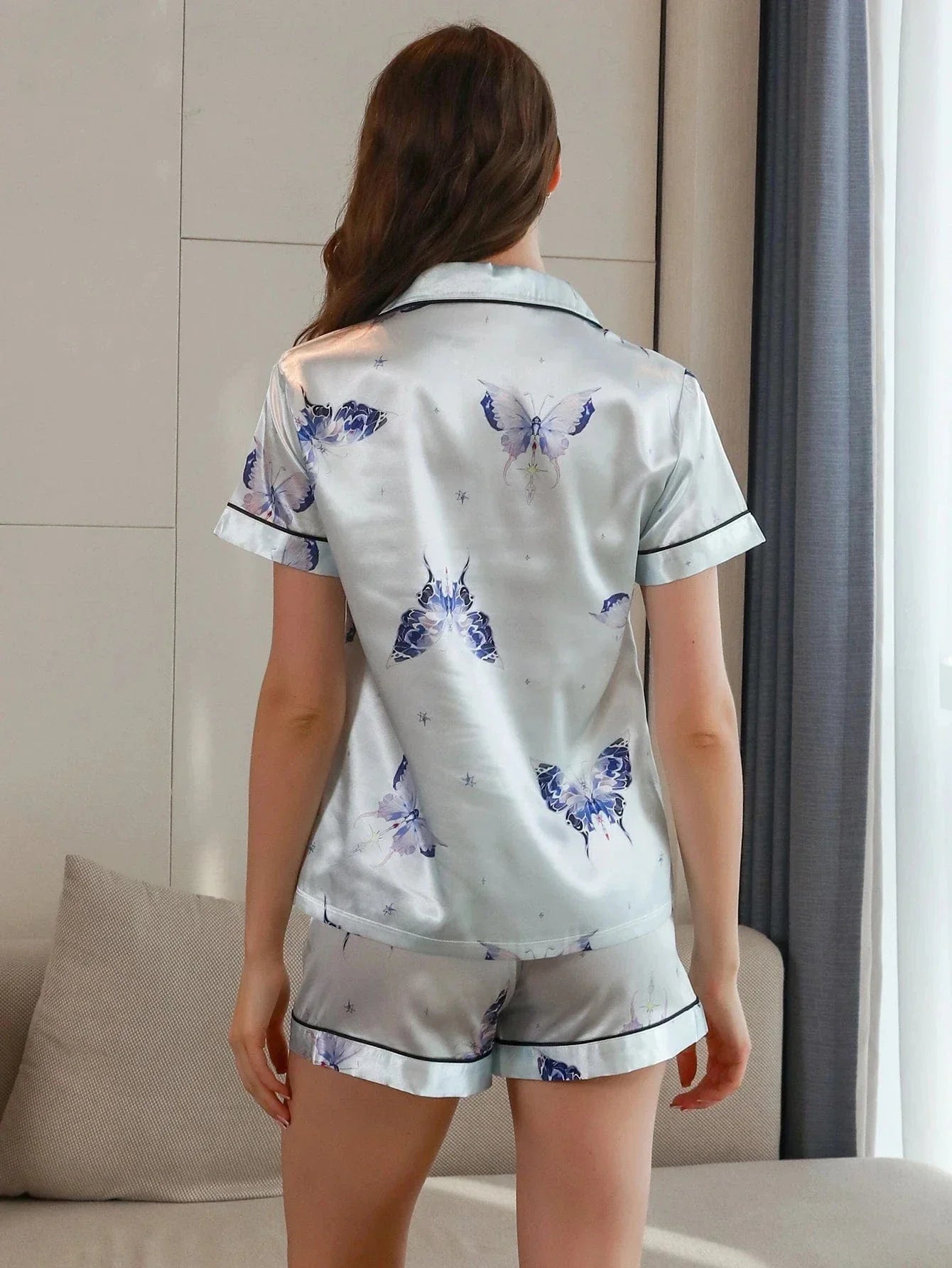 Vue de dos d’une femme portant un pyjama court en satin gris perle, composé d’un haut à manches courtes et d’un short assorti, tous deux ornés de motifs papillons bleus et de passepoils noirs. Ensemble doux, fluide et élégant, idéal pour les nuits confortables ou les moments de détente chic.