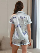 Vue de dos d’une femme portant un pyjama court en satin gris perle, composé d’un haut à manches courtes et d’un short assorti, tous deux ornés de motifs papillons bleus et de passepoils noirs. Ensemble doux, fluide et élégant, idéal pour les nuits confortables ou les moments de détente chic.