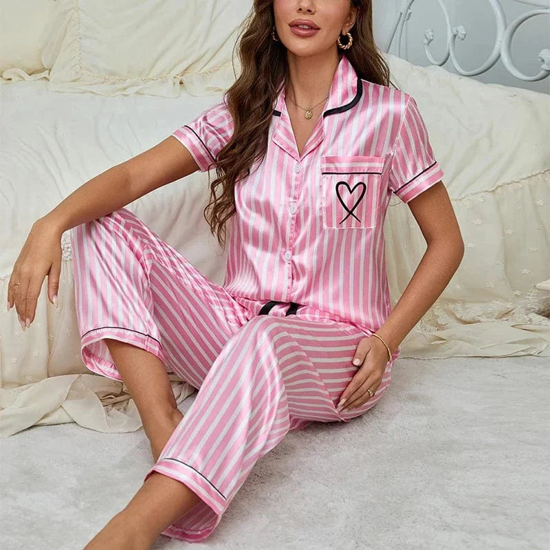 Pyjama en satin pour femme, avec une chemise à rayures roses et blanches, ornée d'une poche décorée d'un cœur. Le pantalon assorti complète ce look à la fois élégant et confortable. Sur cette photo, une femme porte ce pyjama, assise sur un lit dans un cadre cosy, affichant un sourire détendu. Ce pyjama est parfait pour allier confort et style lors de vos moments de détente à la maison.