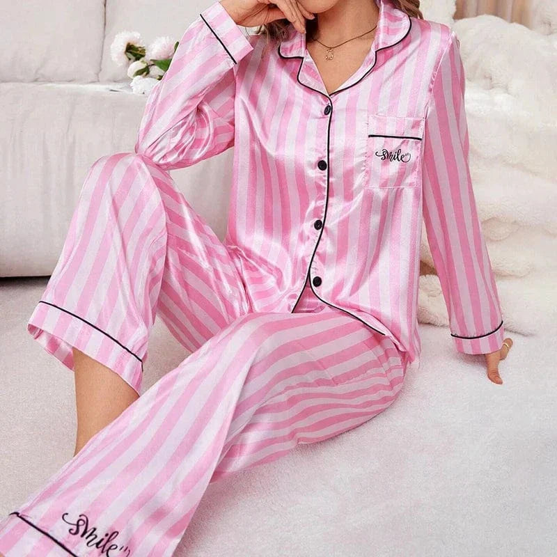 Pyjama en satin pour femme, avec une chemise à rayures roses et blanches et une poche ornée du mot "Smile". Le pantalon assorti complète ce look à la fois chic et confortable. Sur cette photo, une femme porte le pyjama, assise dans un cadre cosy, avec un sourire léger et une pose décontractée. Ce pyjama est parfait pour allier confort et élégance, idéal pour des moments de détente à la maison.
