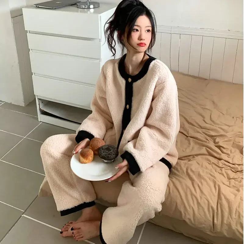Femme assise au bord d’un lit, portant un pyjama polaire beige avec finitions noires contrastantes. Ensemble deux pièces boutonné, doux et épais, parfait pour l’hiver. Elle tient une assiette de viennoiseries, créant une ambiance cosy et gourmande.