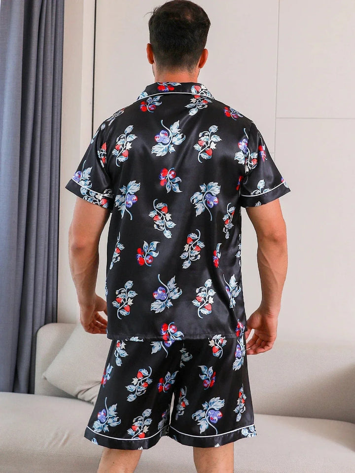 Vue arrière d’un homme portant le pyjama satin Armand pour homme, ensemble deux pièces avec chemise à manches courtes et short assorti, coloris noir à imprimé floral et fruits rouges, finitions contrastées et tissu à l’aspect satiné, présenté en intérieur.