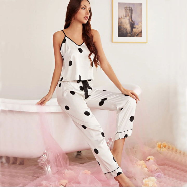 Femme assise au bord d’une baignoire, portant un pyjama satiné blanc à pois noirs, composé d’un débardeur à fines bretelles noires et d’un pantalon fluide avec taille élastique et nœud noir. Ensemble élégant et confortable, parfait pour des soirées détente avec une touche de raffinement.