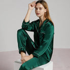 Femme assise en pyjama en satin vert émeraude à manches longues, col à revers et passepoils blancs. Ensemble élégant, fluide et confortable, parfait pour les nuits douces ou les moments de détente raffinés à la maison.
