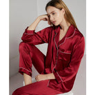 Femme assise en pyjama en satin rouge bordeaux, coupe classique à manches longues avec passepoils blancs et col à revers. Ensemble raffiné et soyeux, offrant douceur et élégance pour des nuits confortables ou des instants de détente chic à la maison.