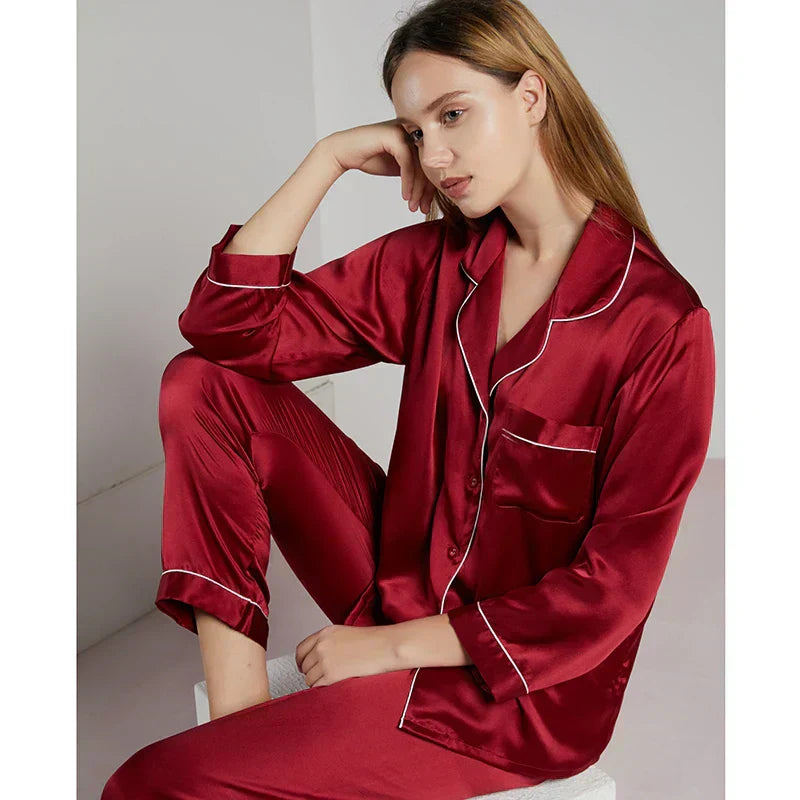 Femme assise en pyjama en satin rouge bordeaux, coupe classique à manches longues avec passepoils blancs et col à revers. Ensemble raffiné et soyeux, offrant douceur et élégance pour des nuits confortables ou des instants de détente chic à la maison.