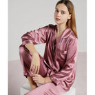 Femme assise portant un pyjama en satin vieux rose, coupe classique avec col à revers, manches longues et passepoils contrastants. Ensemble doux, brillant et raffiné, parfait pour les nuits reposantes ou les moments de détente élégants à la maison.