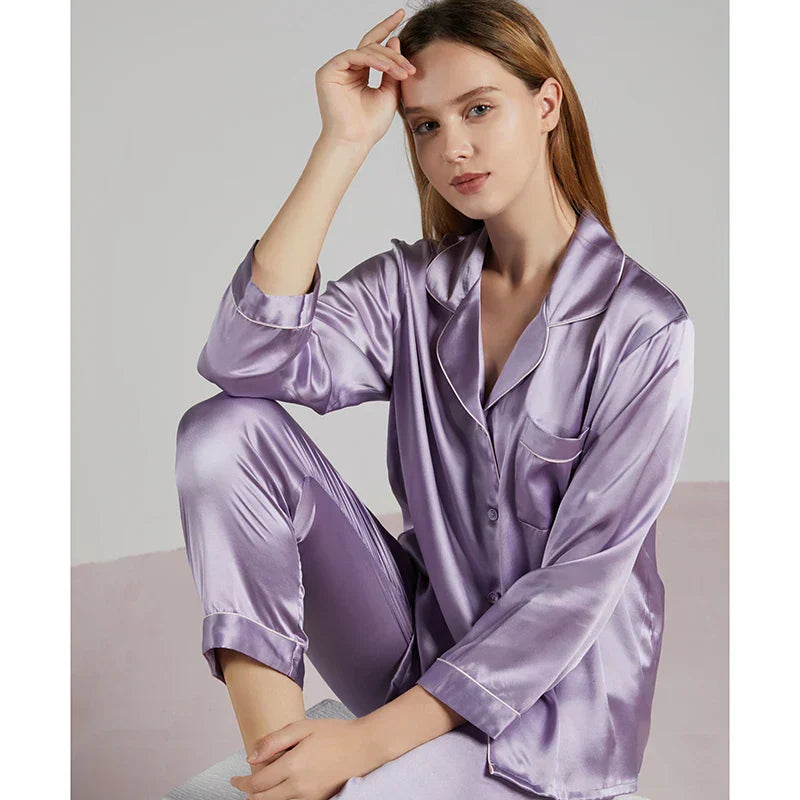Femme assise portant un pyjama en satin violet lavande, manches longues, col à revers et finitions passepoilées. Ensemble raffiné et fluide, idéal pour les nuits paisibles et les instants de détente avec style à la maison.