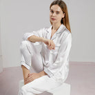 Femme assise en pyjama en satin blanc brillant, à manches longues et col à revers avec passepoils noirs contrastants. Ensemble raffiné et confortable, idéal pour les nuits paisibles et les instants cocooning élégants à la maison.