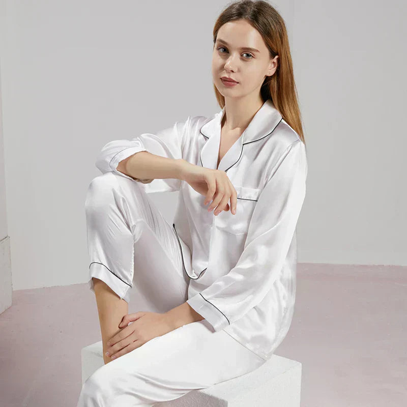 Femme assise en pyjama en satin blanc brillant, à manches longues et col à revers avec passepoils noirs contrastants. Ensemble raffiné et confortable, idéal pour les nuits paisibles et les instants cocooning élégants à la maison.