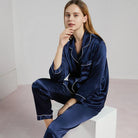 Femme assise portant un pyjama en satin bleu marine à manches longues, col à revers et finitions passepoilées blanches. Ensemble chic et confortable, idéal pour la nuit ou les moments de détente élégants à la maison.