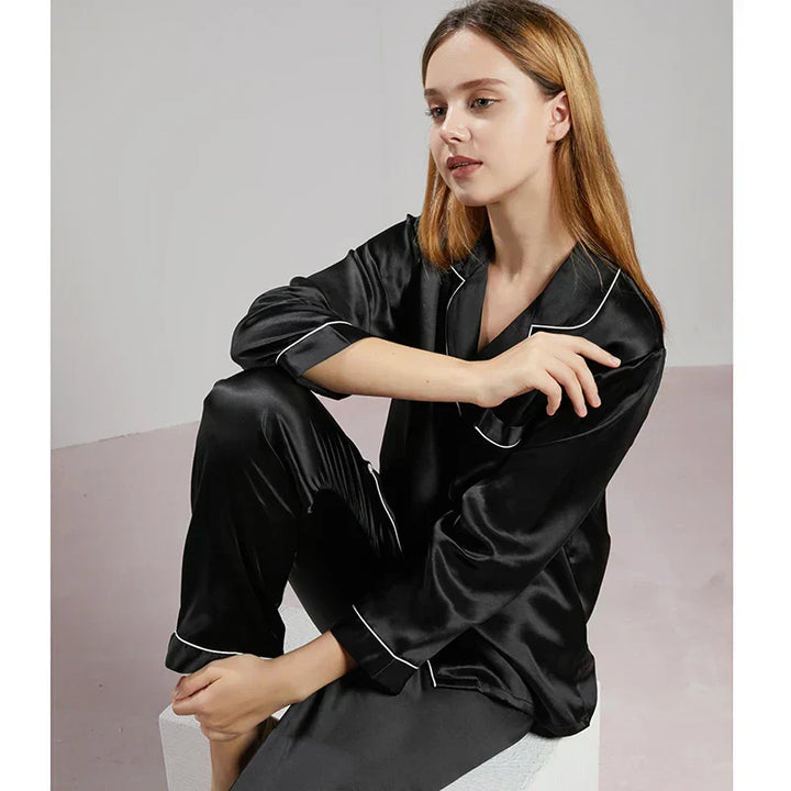 Femme assise en pyjama en satin noir à manches longues, avec col à revers et passepoils blancs. Ensemble raffiné, fluide et brillant, offrant douceur et élégance pour les soirées cocooning ou les nuits confortables à la maison.