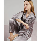 Femme assise portant un pyjama en satin gris perle brillant, à manches longues, col à revers et coupe classique. Ensemble doux et raffiné, idéal pour une nuit confortable ou une routine détente chic à la maison.