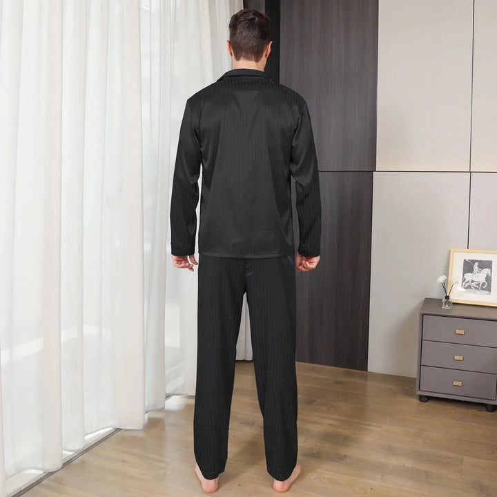 Homme portant le pyjama satin Aymeric pour homme, ensemble noir élégant composé d’une chemise à manches longues et d’un pantalon assorti, coupe droite avec col classique, présenté debout pieds nus dans un intérieur moderne, vue de dos.