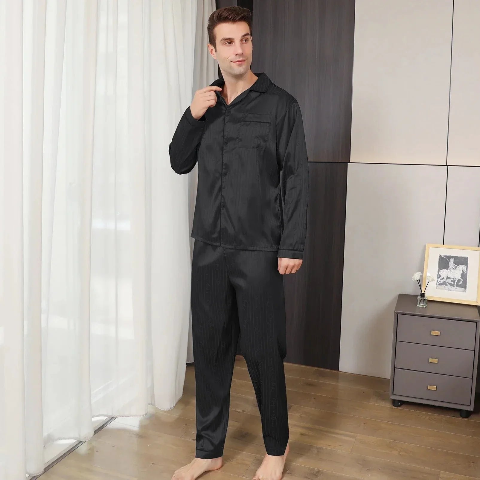 Homme portant le pyjama satin Aymeric pour homme, ensemble noir élégant avec chemise à manches longues et pantalon assorti, col classique et coupe droite, présenté debout pieds nus dans un intérieur moderne, vue de face légèrement de profil.