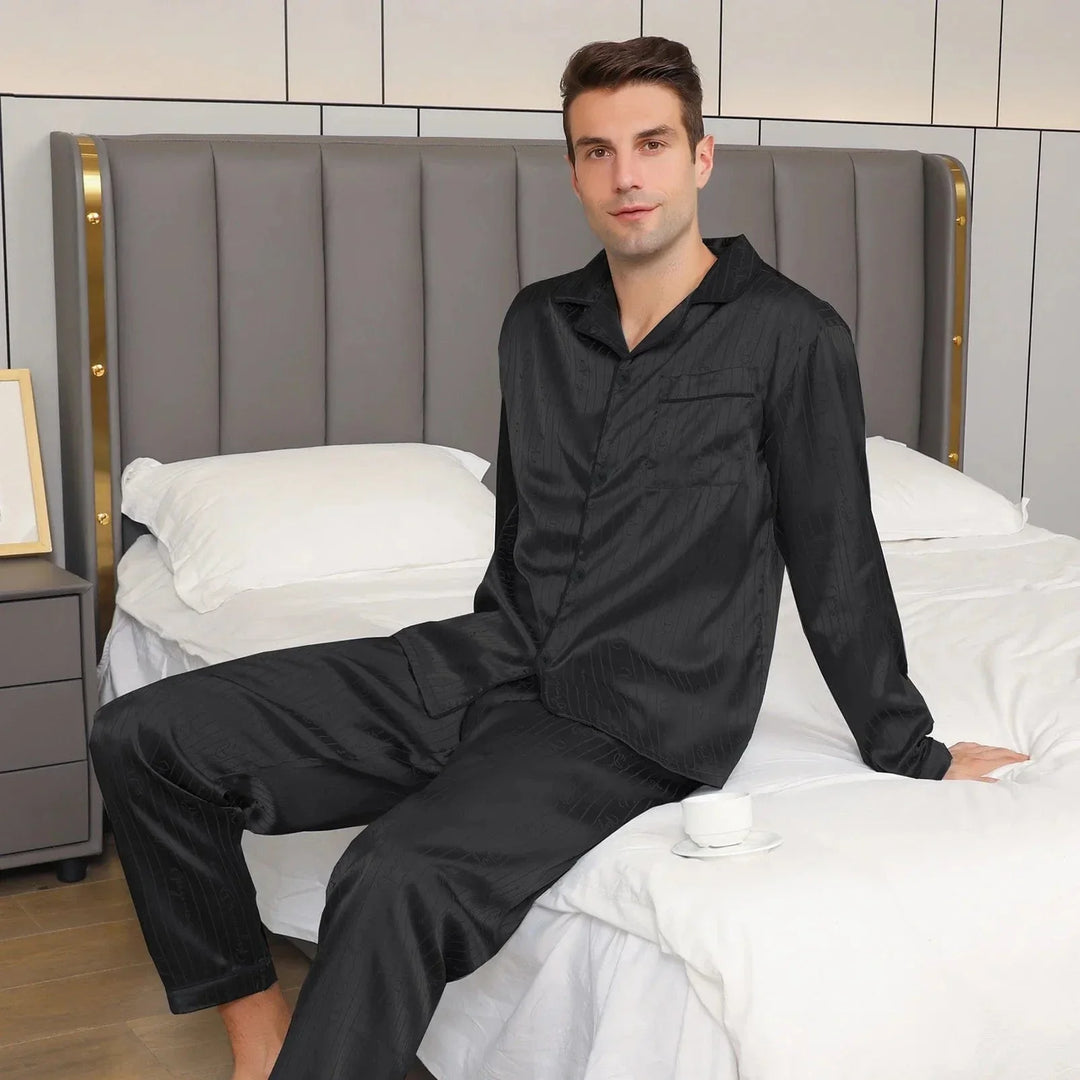 Homme portant le pyjama satin homme Aymeric noir, élégant et confortable, composé d’une chemise à manches longues et d’un pantalon assorti, assis sur un lit dans une chambre moderne.
