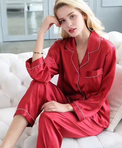 Femme assise sur un canapé, portant un pyjama en satin rouge vif, composé d’une chemise boutonnée à manches longues avec col à revers, poche poitrine et passepoils blancs, accompagnée d’un pantalon fluide assorti. Ensemble chic et confortable, parfait pour les soirées élégantes à la maison.