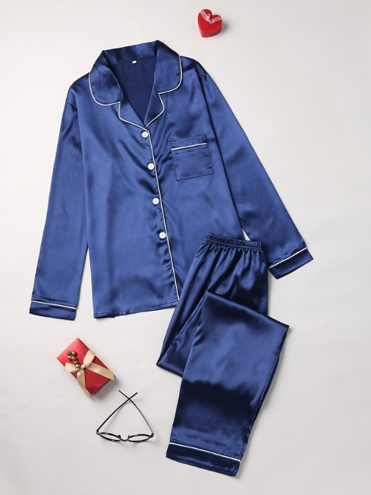 Pyjama femme deux pièces en satin bleu marine avec liserés blancs, composé d'une chemise boutonnée à col à revers et d'un pantalon long assorti, élégamment disposé à plat avec des accessoires de nuit comme des lunettes et un coffret cadeau, créant une ambiance chic et cosy.