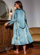 Vue arrière de la femme portant la robe-kimono bleu satinée avec des motifs floraux dorés, les bras légèrement ouverts. Le kimono s'étend jusqu'aux chevilles, créant un look élégant et raffiné. Idéal pour une soirée chic ou un moment de détente avec une touche de luxe.