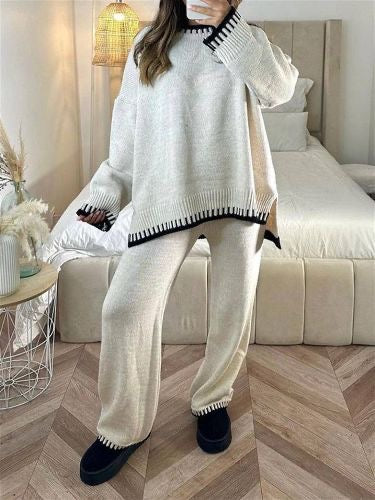 Femme debout dans une chambre moderne, portant un ensemble lounge femme beige avec finitions noires contrastantes sur les manches, le col, l’ourlet du haut et le bas du pantalon. Tenue ample et confortable, au style chic et décontracté, parfaite pour la maison ou les sorties casual.